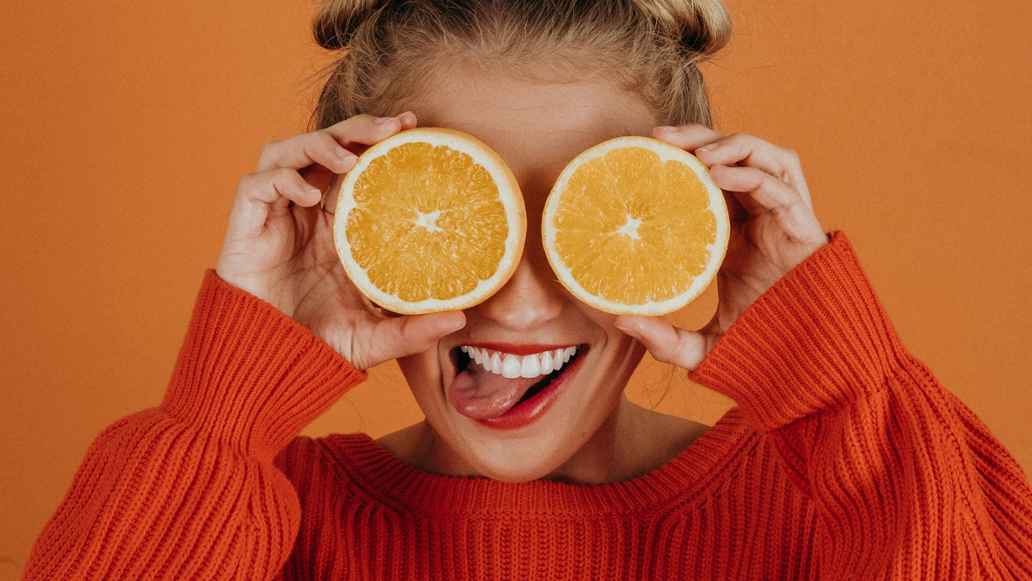 Woman putting oranges in front of her face sticks out her tongue in a playful way.